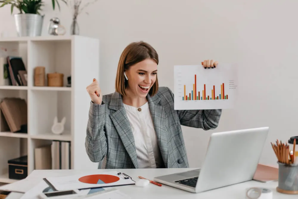 Mulher sorridente vestindo blazer comemora ao exibir um gráfico de barras em frente ao notebook em sua mesa de trabalho.