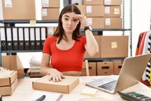 Mulher com caixas de papelão separando produtos da sua loja virtual, com a mão na cabeça, indicando esquecimento