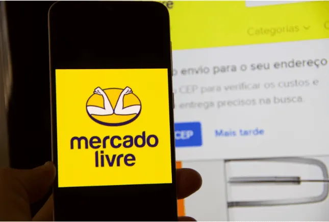 Mão segurando um smartphone que exibe o logotipo do Mercado Livre na tela, com a página do site da empresa aberta em um computador ao fundo.
