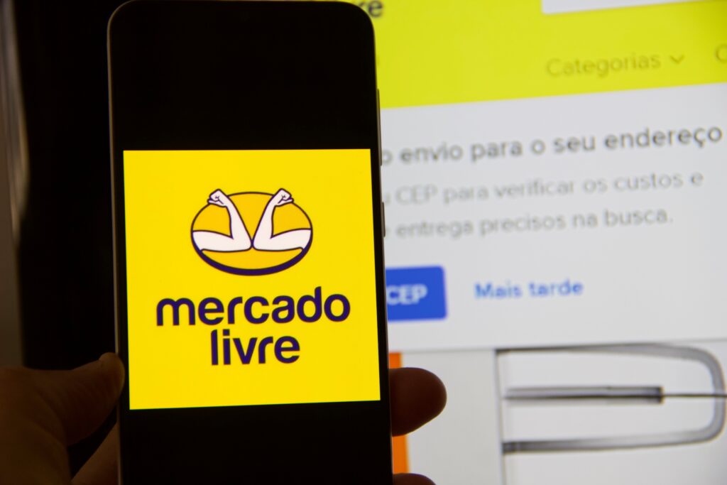 Celular com logo do Mercado Livre e página do computador aberta atrás.