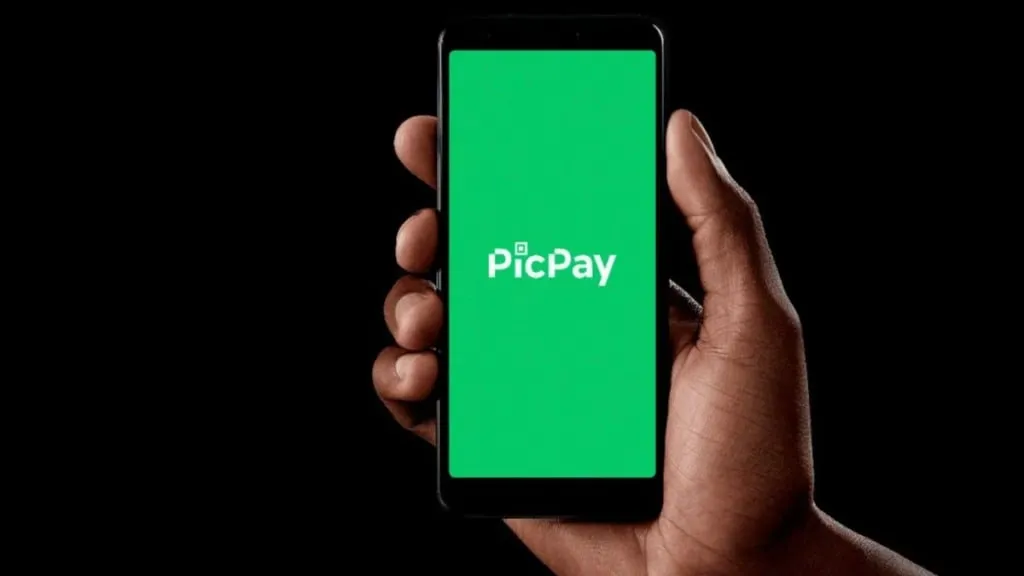 Uma mão segura um smartphone com a tela exibindo o logotipo do PicPay em fundo verde, sobre um fundo preto
