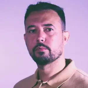 Homem de meia-idade com cabelos e barba grisalhos, usando óculos de grau de armação fina e blusa de frio escura, olhando para a câmera com expressão serena.