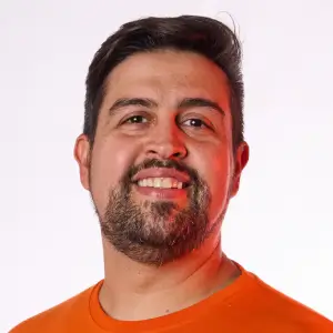 Homem de meia-idade com cabelos e barba grisalhos, usando óculos de grau de armação fina e blusa de frio escura, olhando para a câmera com expressão serena.
