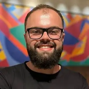 Homem de meia-idade com cabelos e barba grisalhos, usando óculos de grau de armação fina e blusa de frio escura, olhando para a câmera com expressão serena.