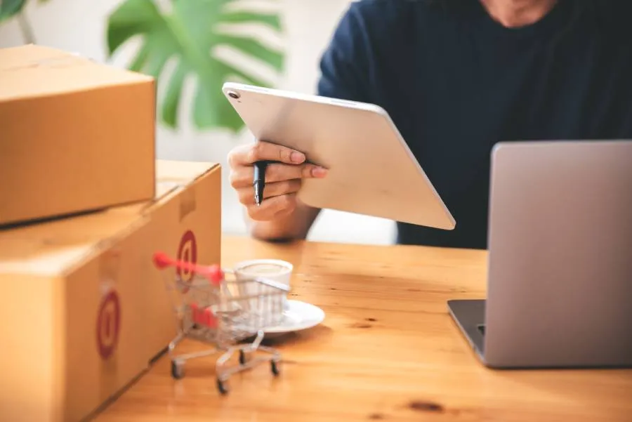 Seller de marketplace organizando pedidos e analisando vendas em tablet, com caixas de envio ao lado.