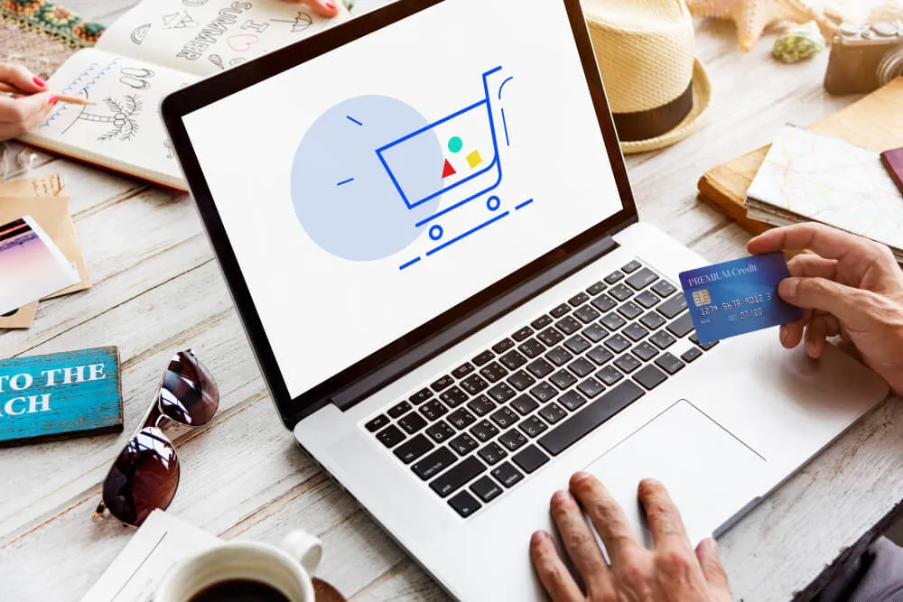 Computador com imagem de carrinho simbolizando compra em loja virtual e pessoa com cartão de crédito na mão