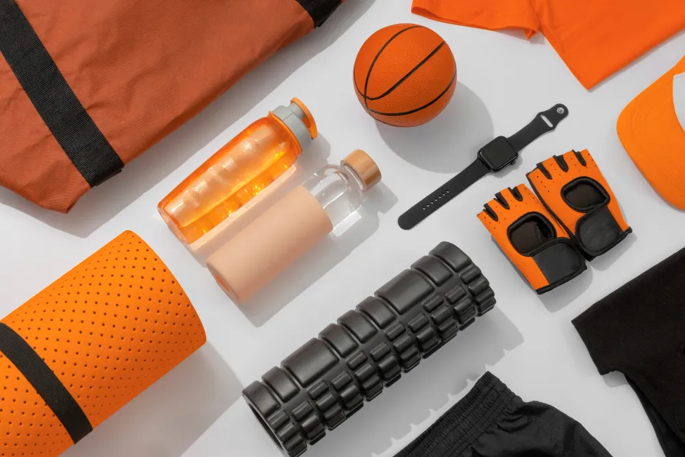 Equipamentos esportivos e acessórios de treino nas cores laranja e preto dispostos sobre um fundo branco em estilo flat lay, incluindo tapete de yoga enrolado, rolo de liberação miofascial, garrafas de água, relógio inteligente, luvas de academia e mini bola de basquete.