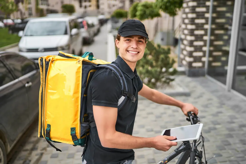 Entregador com mochila de delivery ao lado de bicicleta e segurando um tablet, representando entregas realizadas pelo 99Compras.